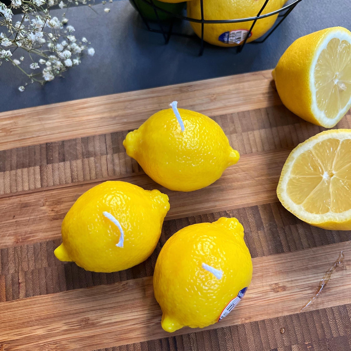 Three handmade lemon-shaped candles styled beside a freshly sliced lemon on a wooden cutting board in a bright, natural setting.