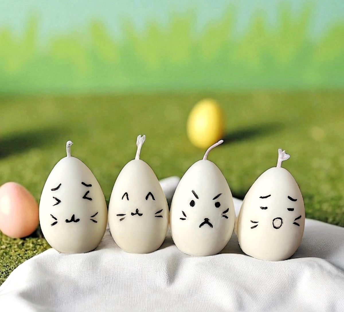 Four handmade egg-shaped candles with cute facial expressions displayed on white fabric with colorful Easter eggs in the background.