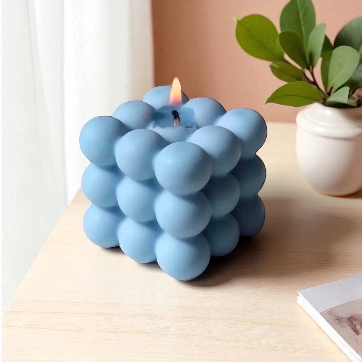 Blue bubble cube soy candle burning on a wooden table beside a white ceramic planter with green leaves.