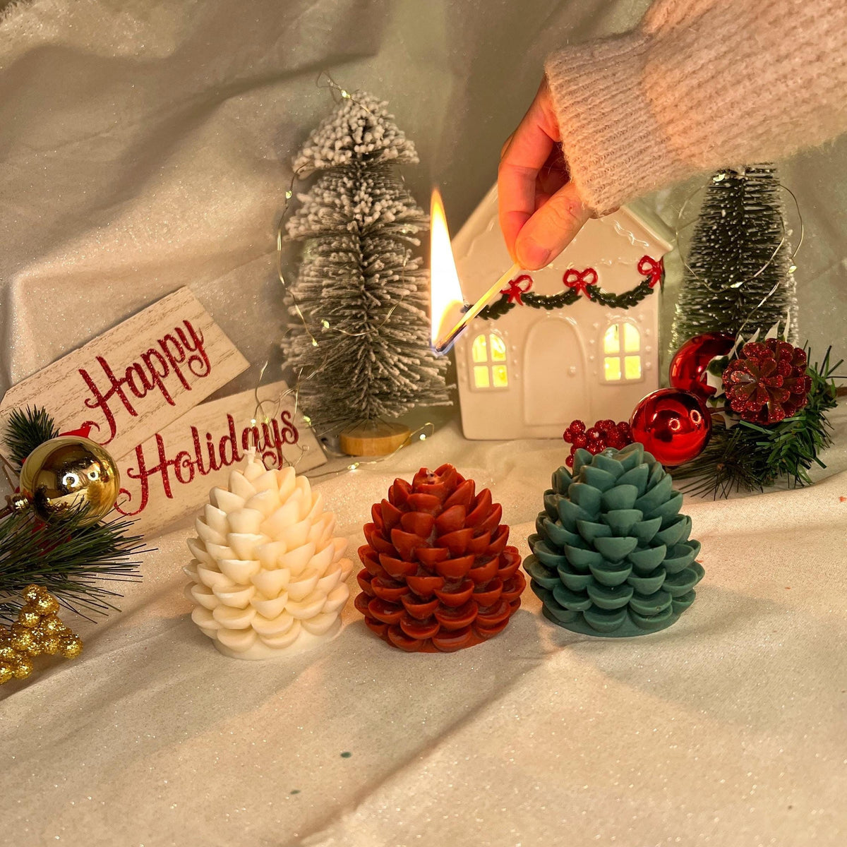 Three handmade Christmas candles in white, red, and green on a festive holiday background as a hand lights one with a burning match.