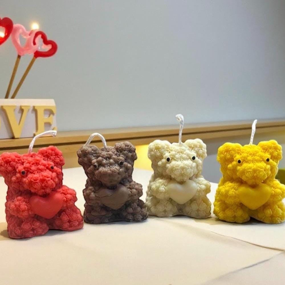 Four handmade teddy bear candles in red, brown, white, and yellow displayed against a romantic heart-themed background.