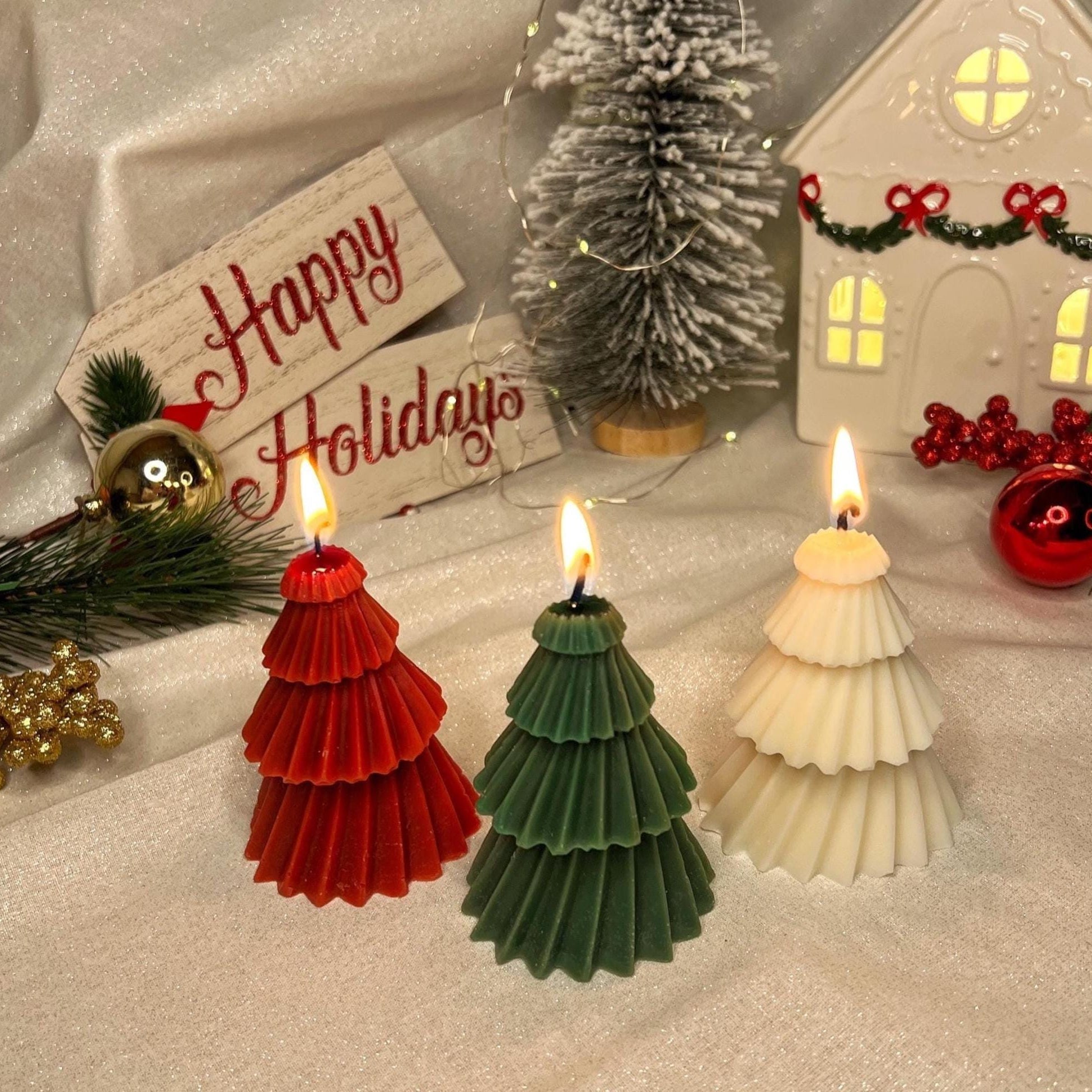 Three Christmas-themed pinecone candles in white, red, and teal are arranged on a white fabric backdrop, with a hand lighting one of the candles. The scene is decorated with miniature Christmas trees, a white house ornament, and a 'Happy Holidays' sign.