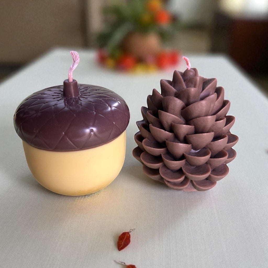 Two scented candles shaped like an acorn and a pinecone, displayed on a light-colored table with a blurred indoor background.