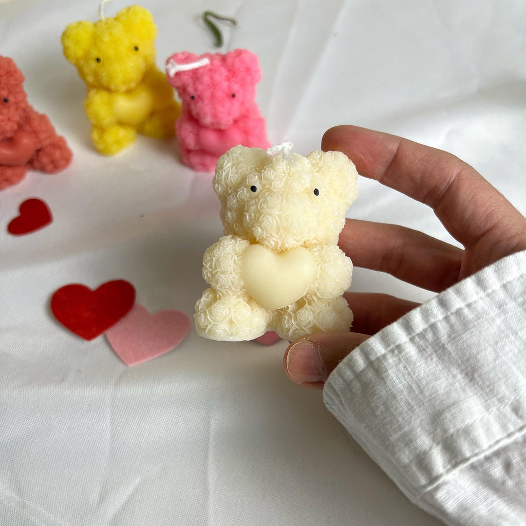 A hand wearing a white long-sleeve shirt holds a small cream-colored teddy bear candle with a heart-shaped center. In the background, other colorful teddy bear candles in yellow, pink, and coral are arranged on a white fabric surface with red and pink heart-shaped decorations.