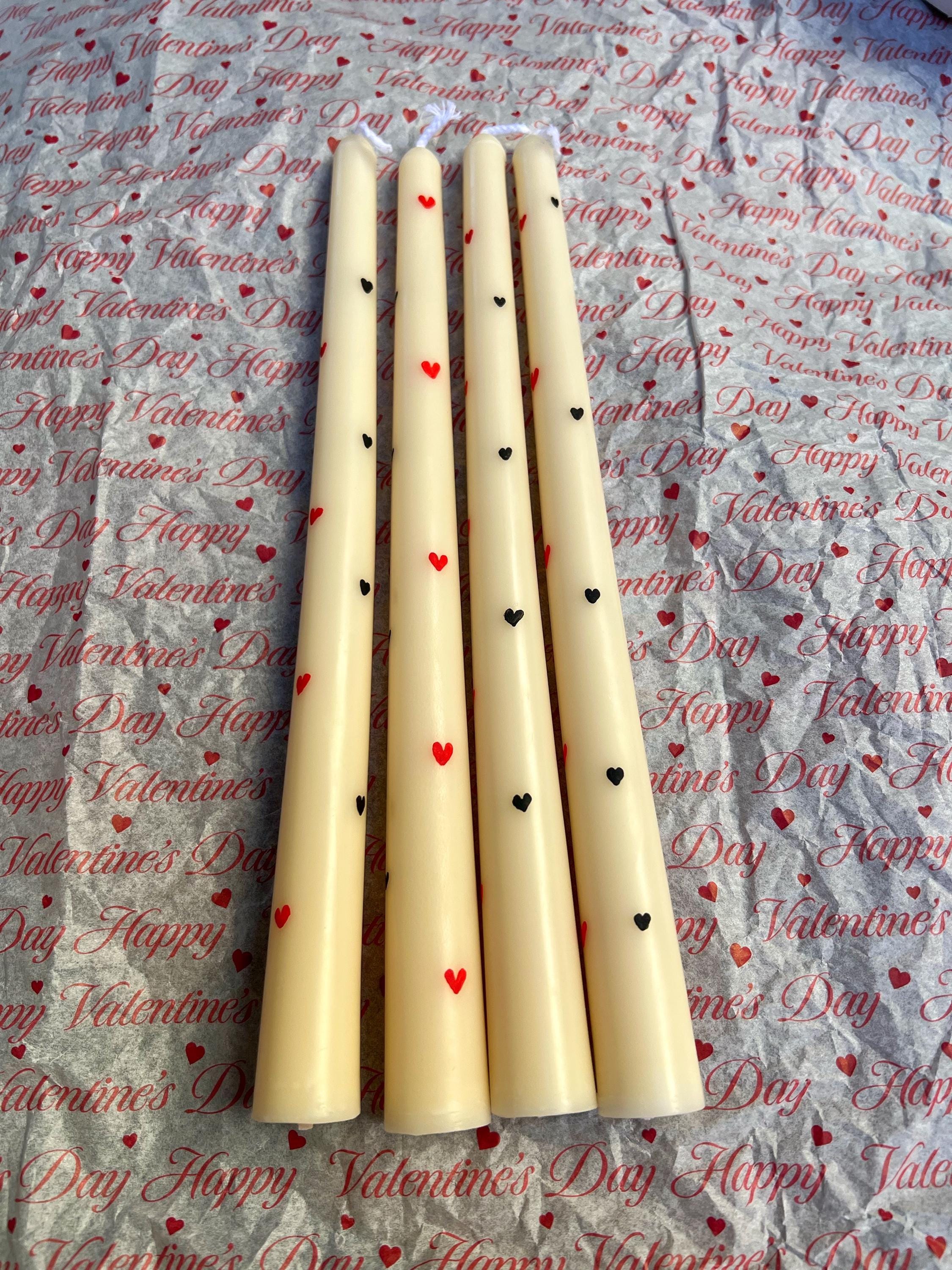 Four cream-colored taper candles decorated with small red and black hearts, arranged vertically on a patterned Valentine's Day fabric background.