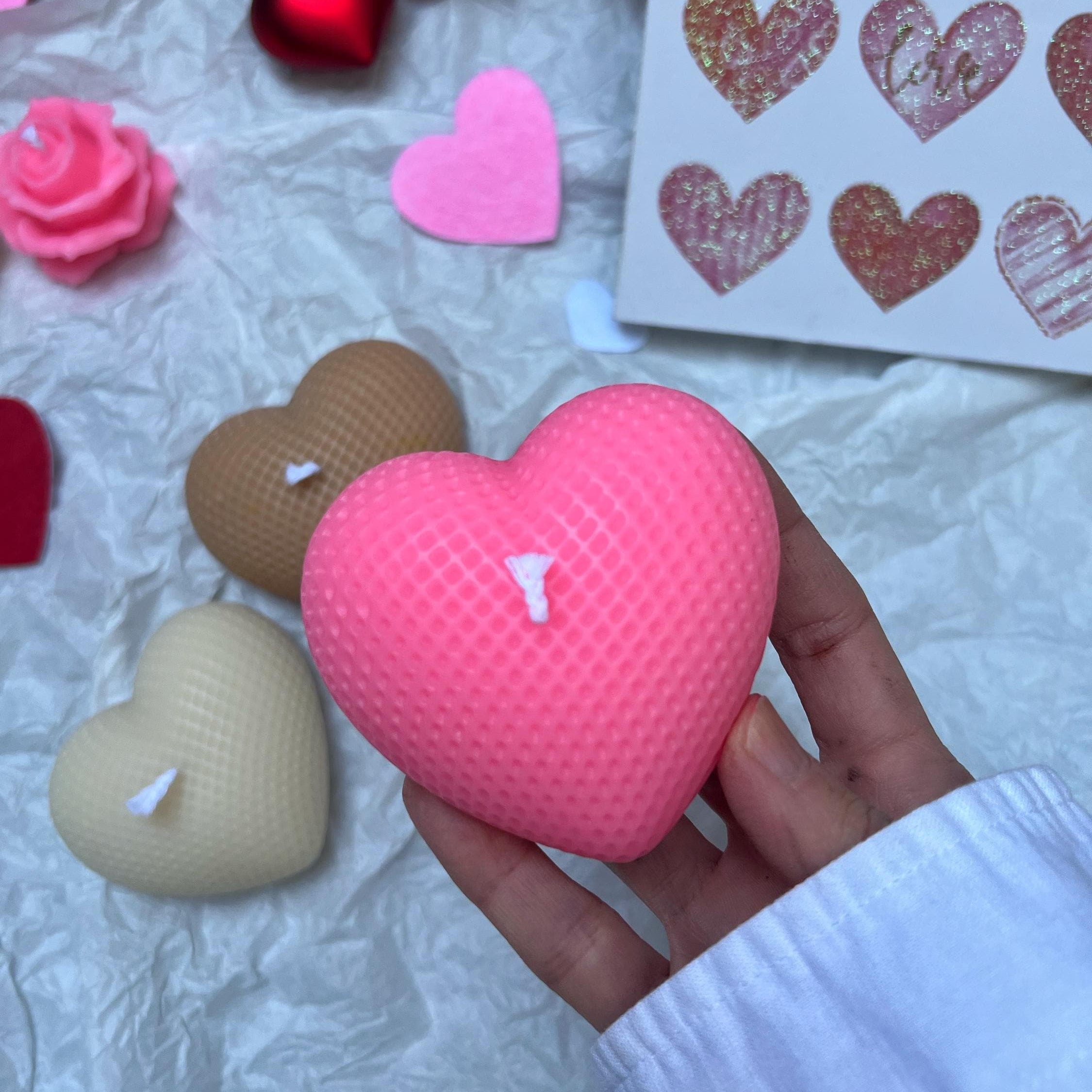 A hand holds a pink heart-shaped textured candle in the foreground, with other heart-shaped candles and a sheet of heart stickers in the background on a crumpled white surface.