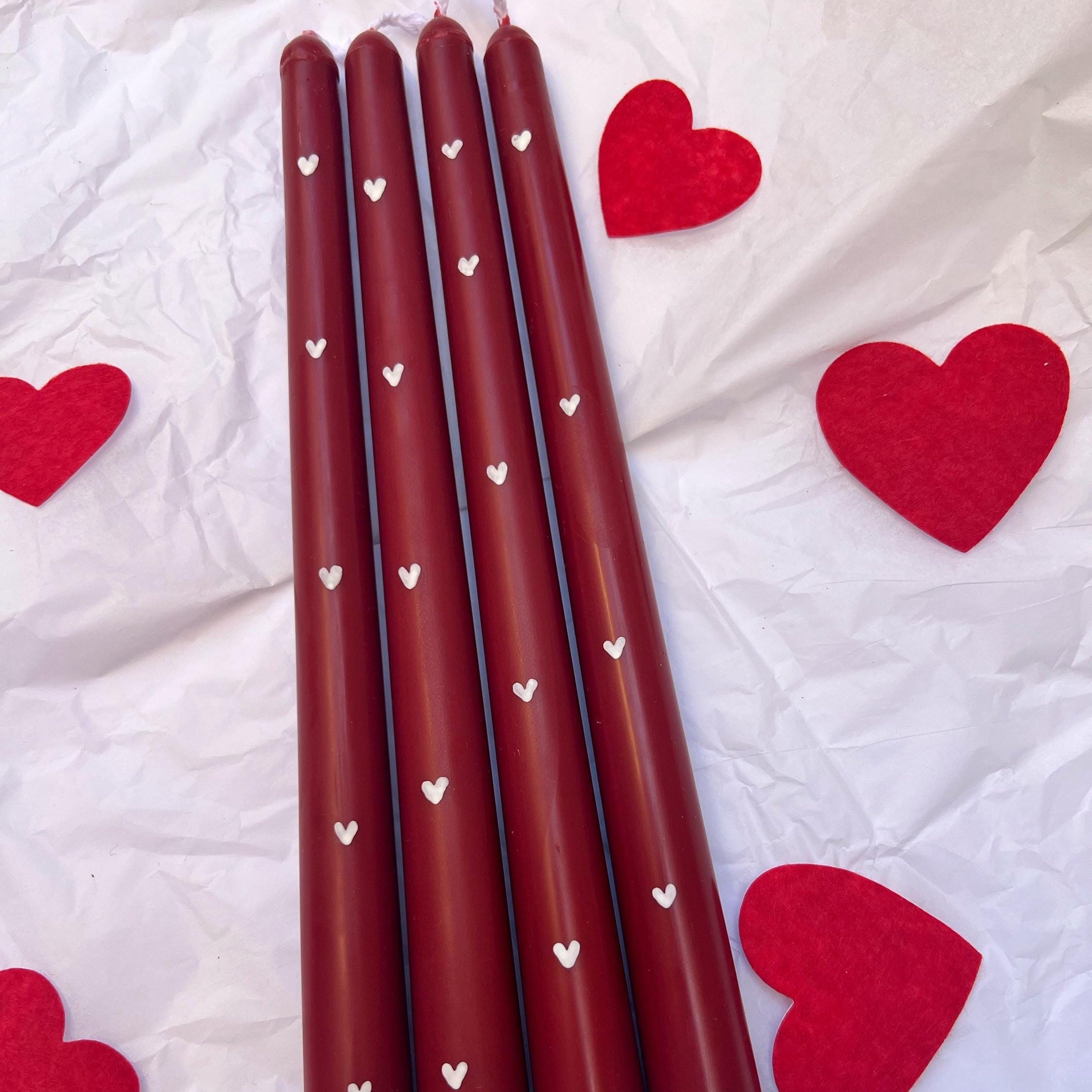 Five dark red taper candles with small white heart patterns, arranged on a crumpled white surface with red felt heart decorations scattered around.