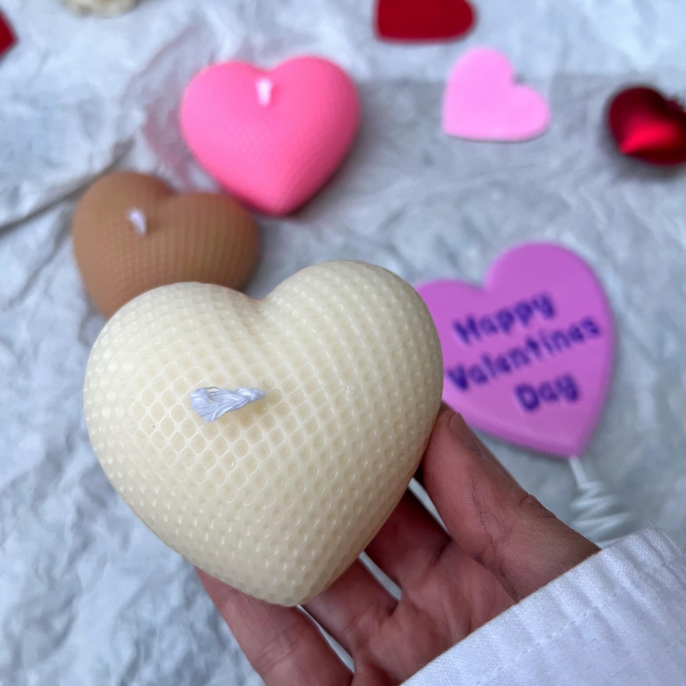 A hand holds a textured cream-colored heart-shaped candle in the foreground, with other heart-shaped candles and a pink 'Happy Valentine's Day' candle scattered on a white fabric background.
