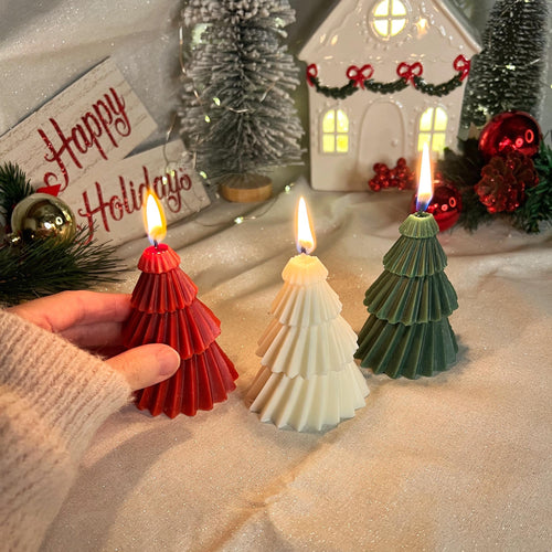 Three Christmas tree-shaped candles are lit on a glittery beige fabric surface, with a hand holding the red candle on the left. The scene is decorated with miniature trees, a white holiday house, ornaments, and a 'Happy Holidays' sign.