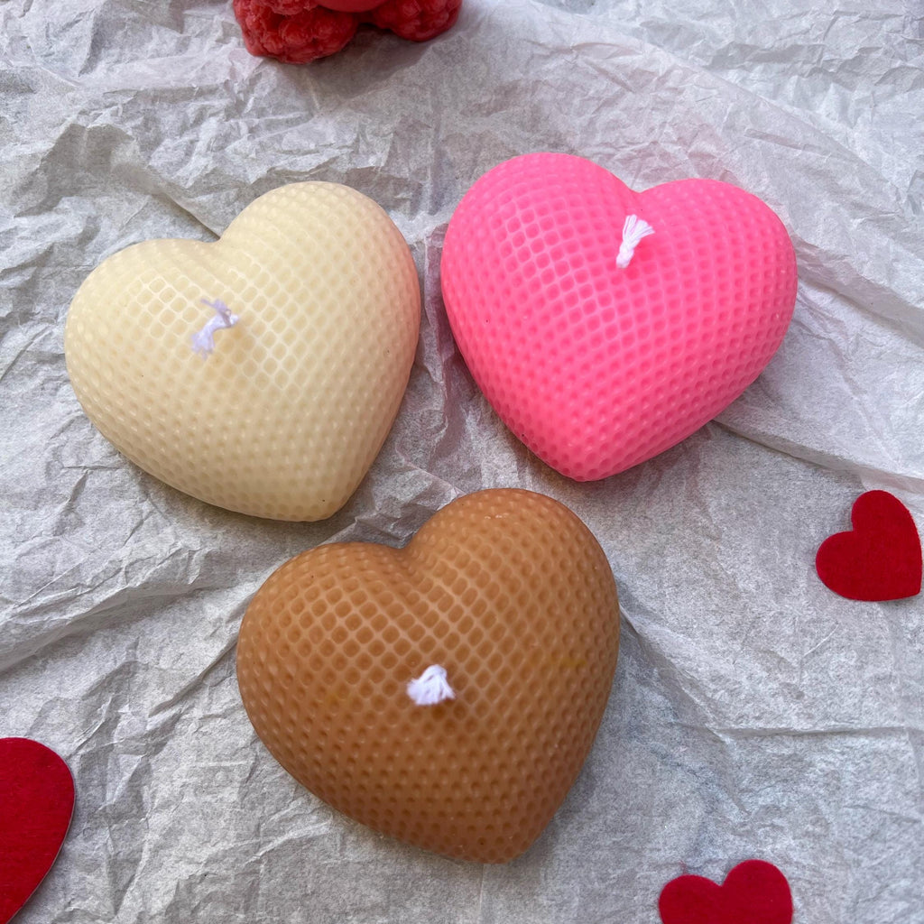 Three heart-shaped textured candles in beige, pink, and brown, arranged on crumpled silver paper with small red heart cutouts and a red teddy bear decoration in the background.