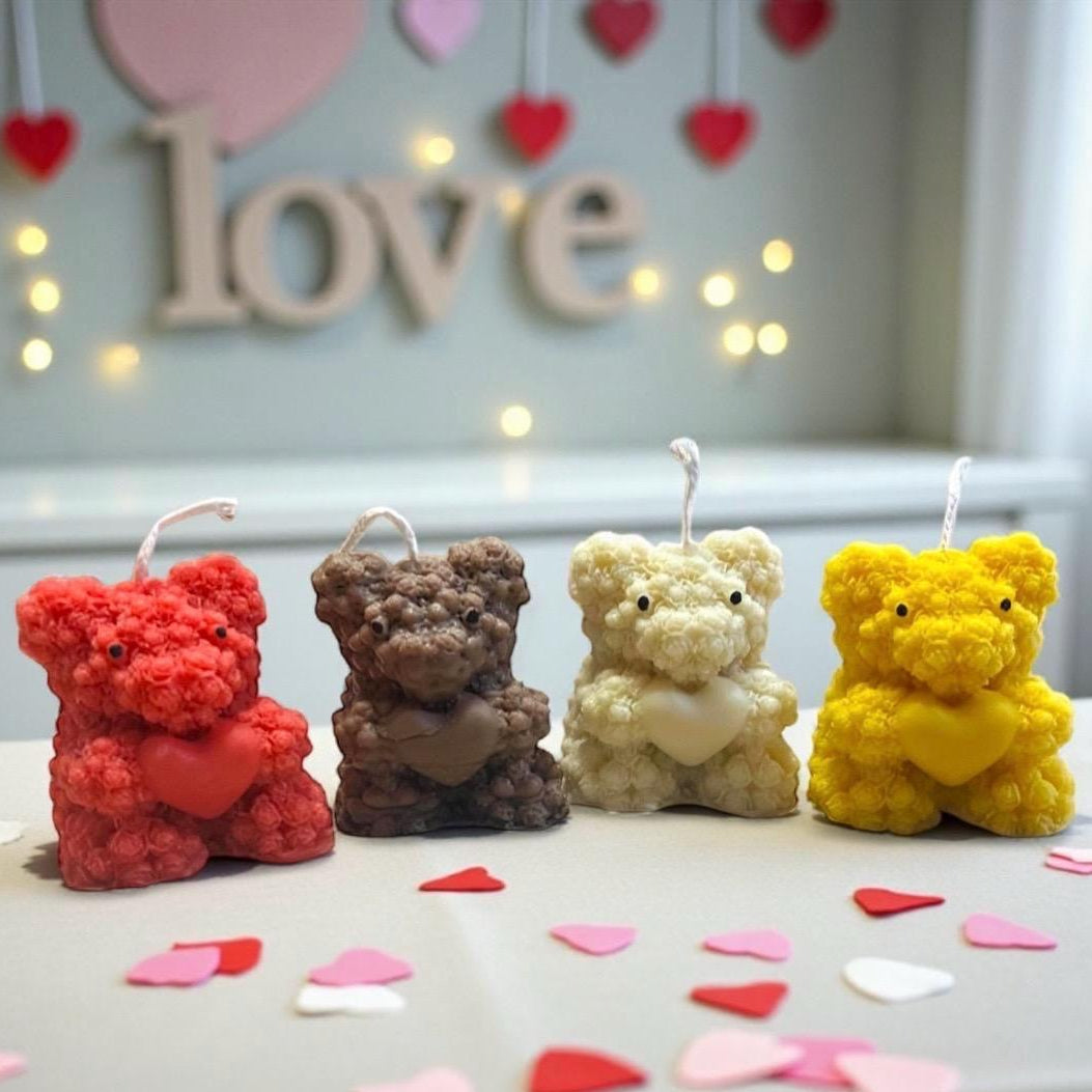 Four teddy bear-shaped candles in red, brown, cream, and yellow, arranged on a table with scattered heart-shaped confetti in the foreground and a blurred 'love' sign in the background.