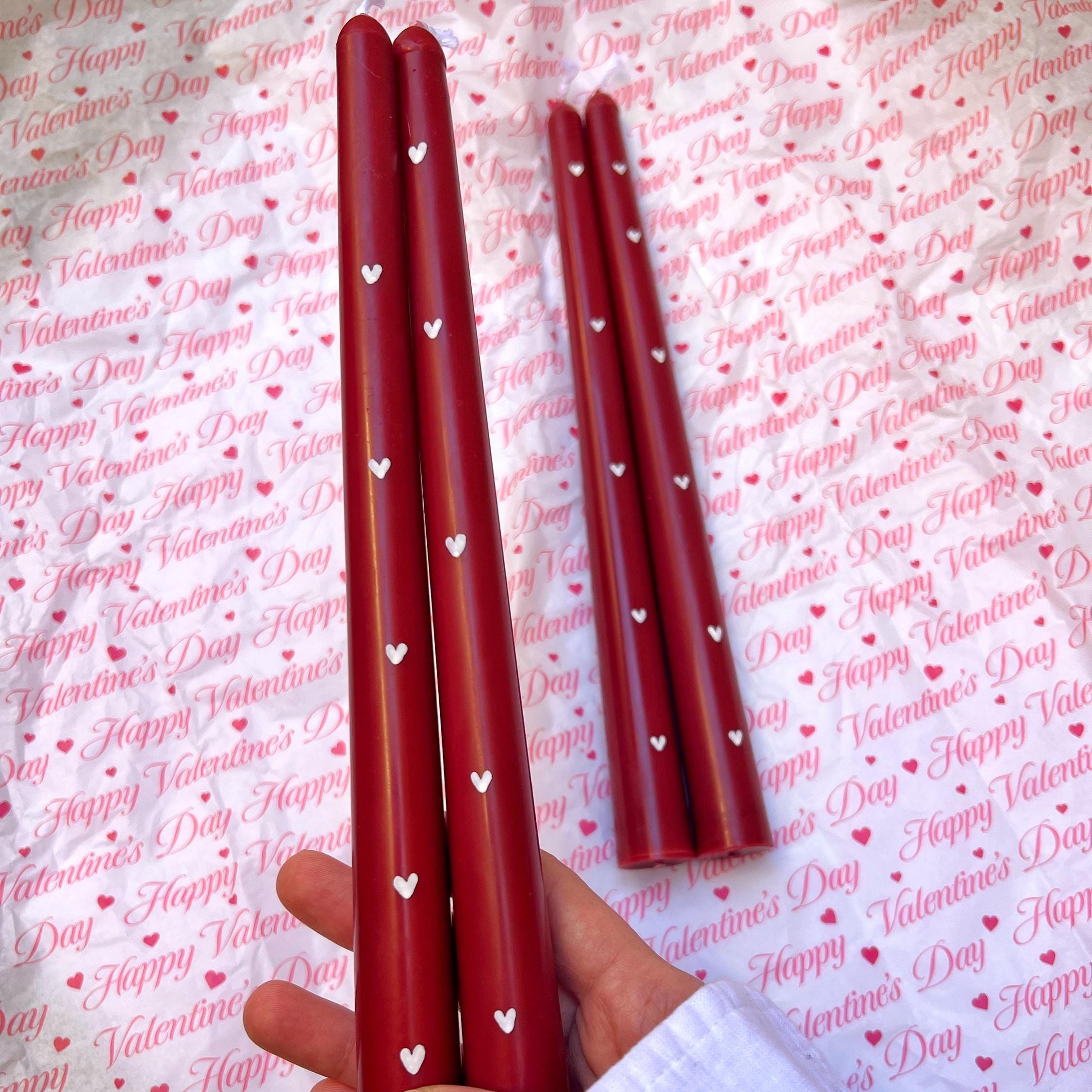 Three red taper candles with small white heart prints are held in a hand, with two more standing upright in the background on a white surface covered in a repeating Valentine's Day pattern.