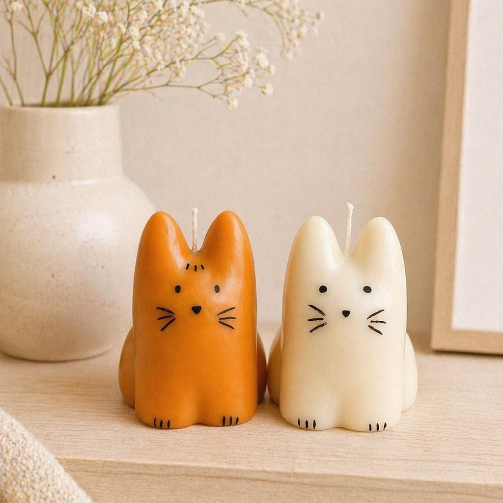 Two cat-shaped candles, one orange and one cream, sitting on a light wood shelf next to a ceramic vase with dried flowers.