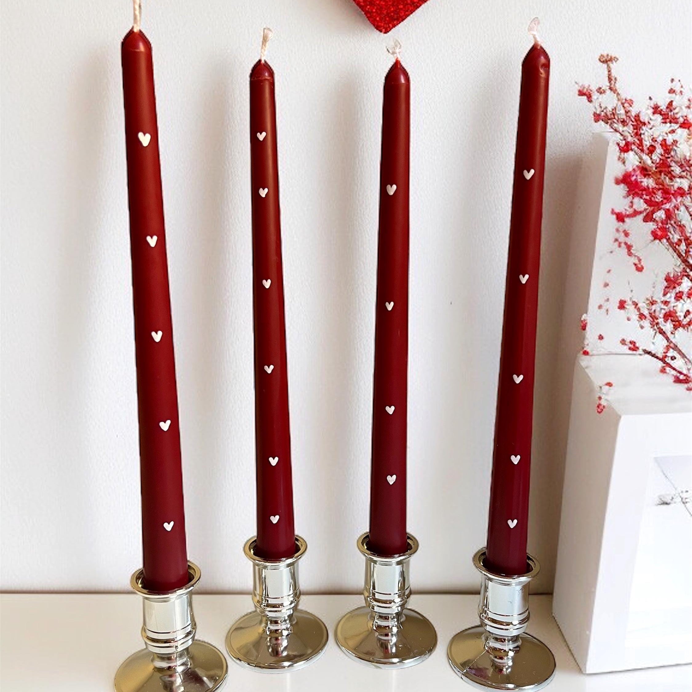 Four tall dark red taper candles with small white heart cutouts, displayed in silver candlesticks on a white surface against a white wall, with a red heart decoration and red floral branches in the background.