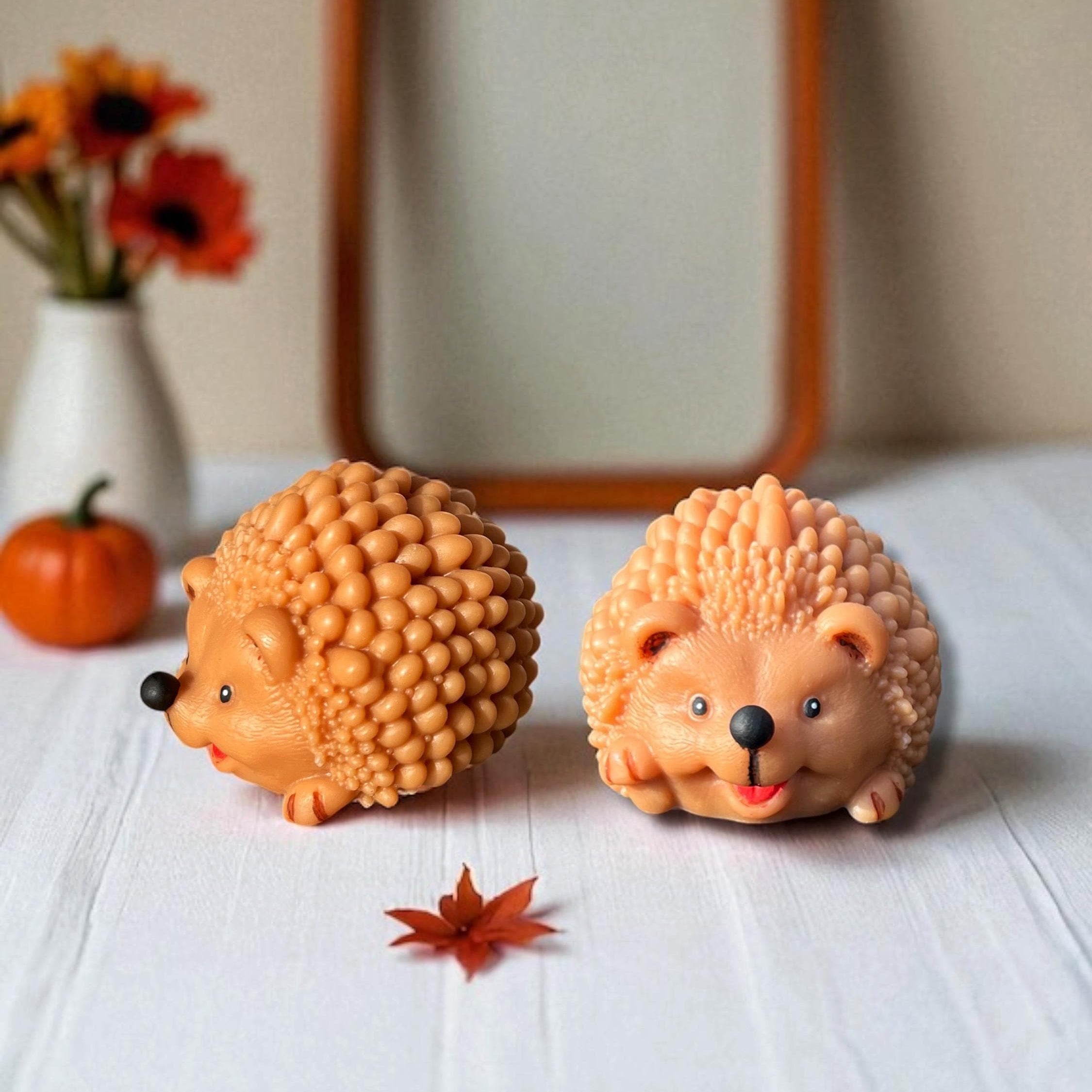 Two small hedgehog figurines in warm peach and orange tones, placed on a white surface with a blurred wooden frame, flowers, and a pumpkin in the background.