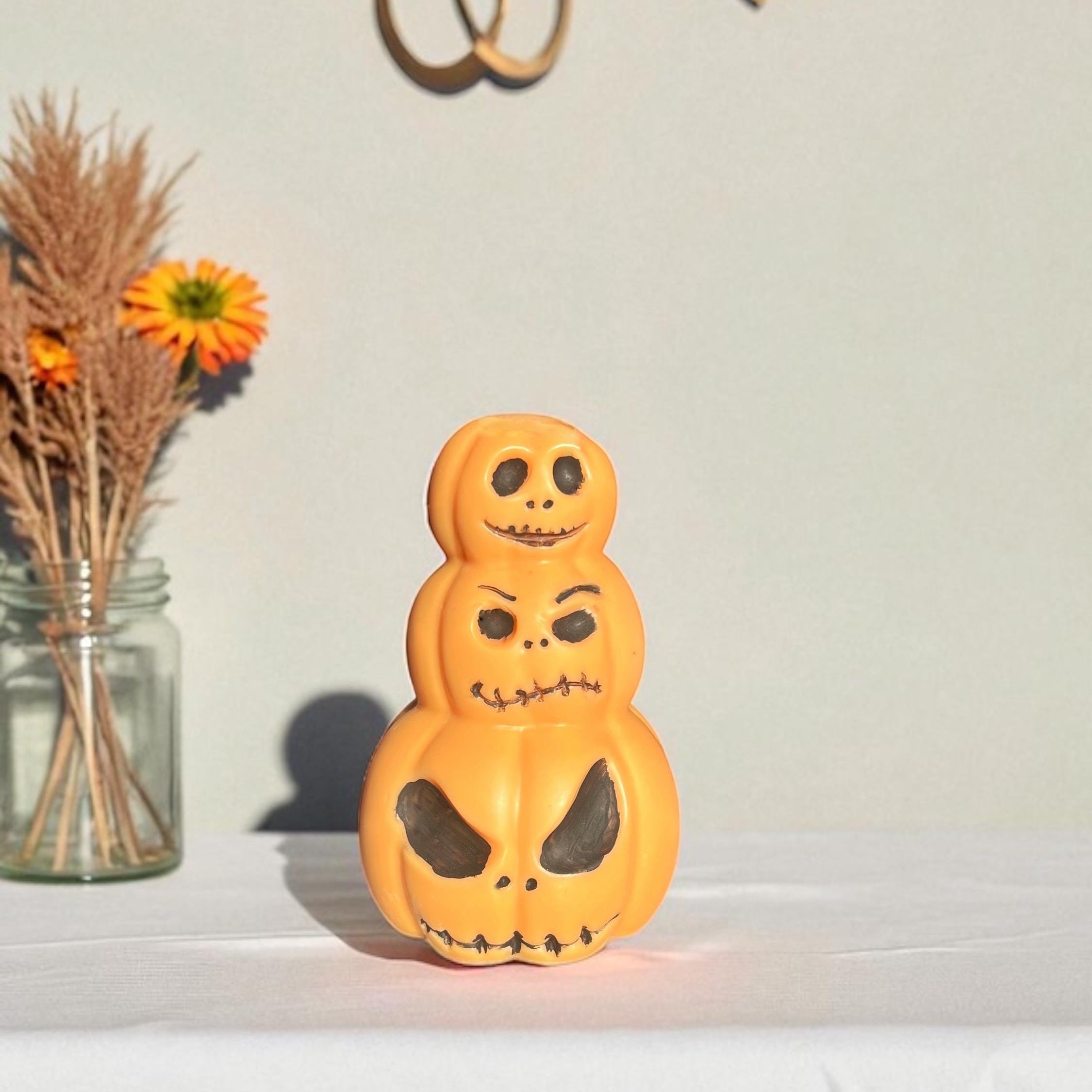 A small orange stacked jack-o'-lantern decoration with carved faces sits on a white draped surface against a light wall, with dried flowers in a glass jar at left.