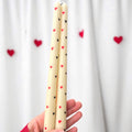A hand holding two cream-colored taper candles decorated with small red and black hearts, set against a white curtain backdrop with blurred red heart decorations.