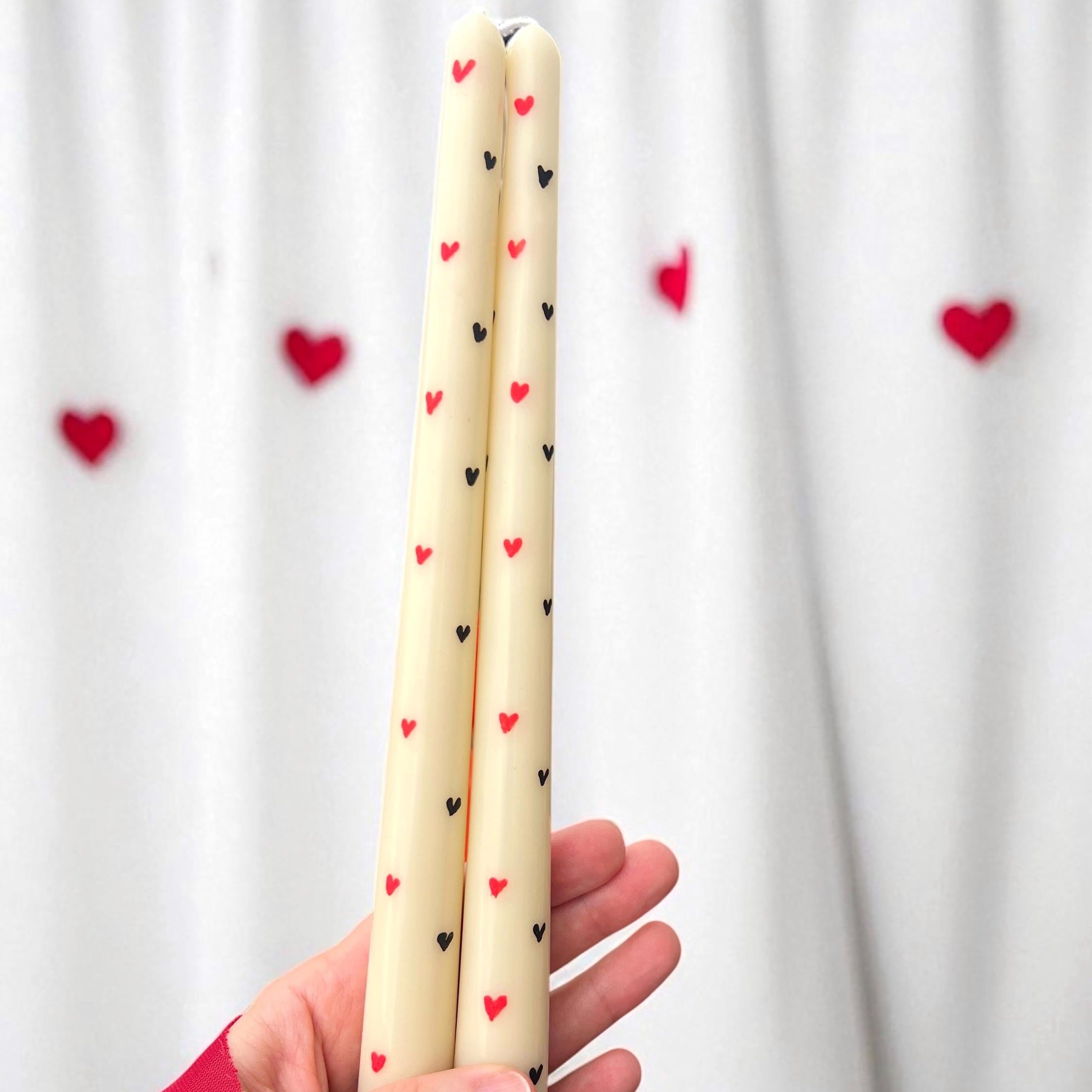 A hand holding two cream-colored taper candles decorated with small red and black hearts, set against a white curtain backdrop with blurred red heart decorations.