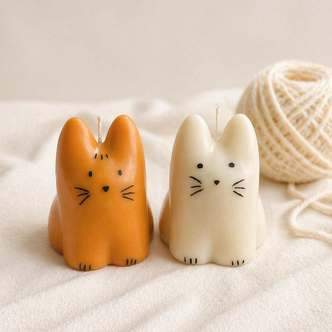 Two small cat-shaped candles, one orange and one cream, with black whisker details, placed on a soft cream fabric next to a ball of cream yarn.