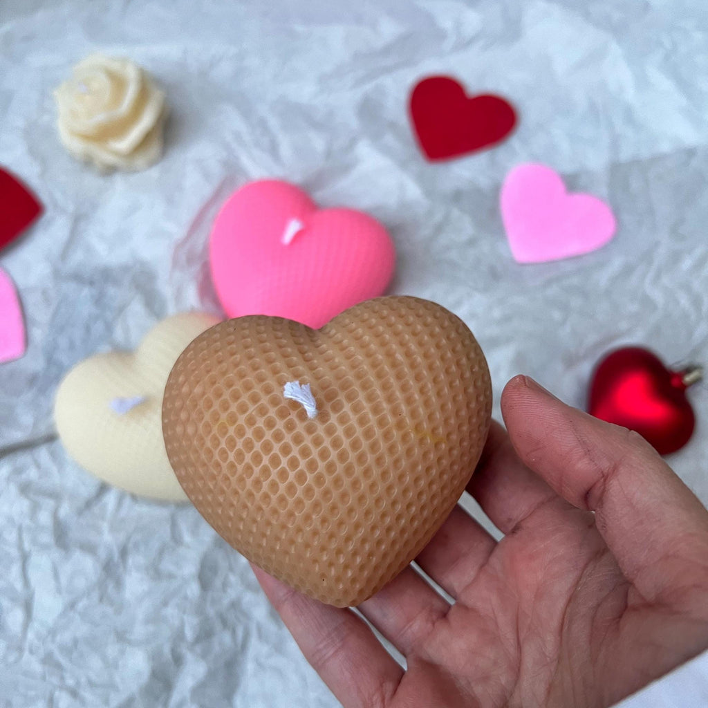 A hand holding a heart-shaped honeycomb candle in a light brown color, with other heart-shaped candles and decorative hearts scattered on a white crumpled paper background.