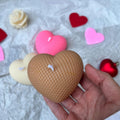A hand holding a heart-shaped honeycomb candle in a light brown color, with other heart-shaped candles and decorative hearts scattered on a white crumpled paper background.