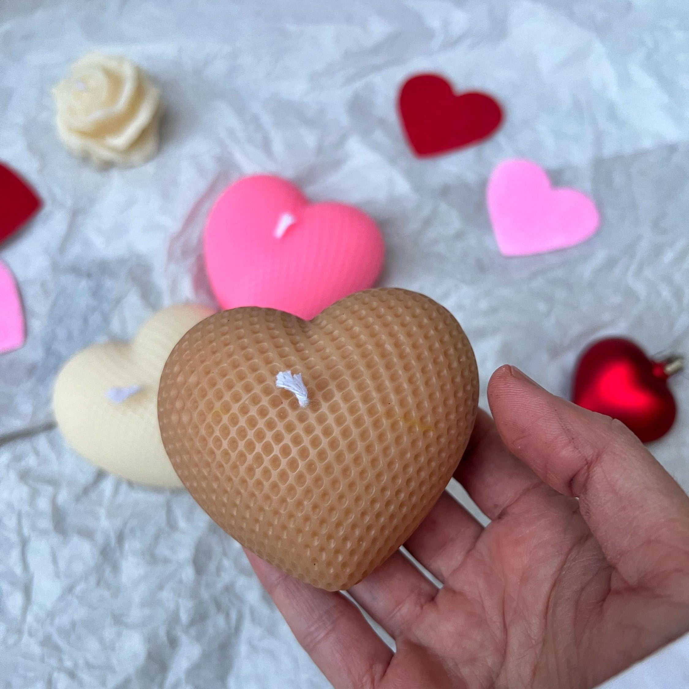 A hand holding a heart-shaped honeycomb candle in a light brown color, with other heart-shaped candles and decorative hearts scattered on a white crumpled paper background.