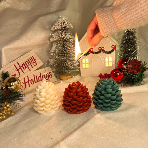 Three Christmas-themed pinecone candles in white, red, and teal are arranged on a white fabric backdrop, with a hand lighting one of the candles. The scene is decorated with miniature Christmas trees, a white house ornament, and a 'Happy Holidays' sign.