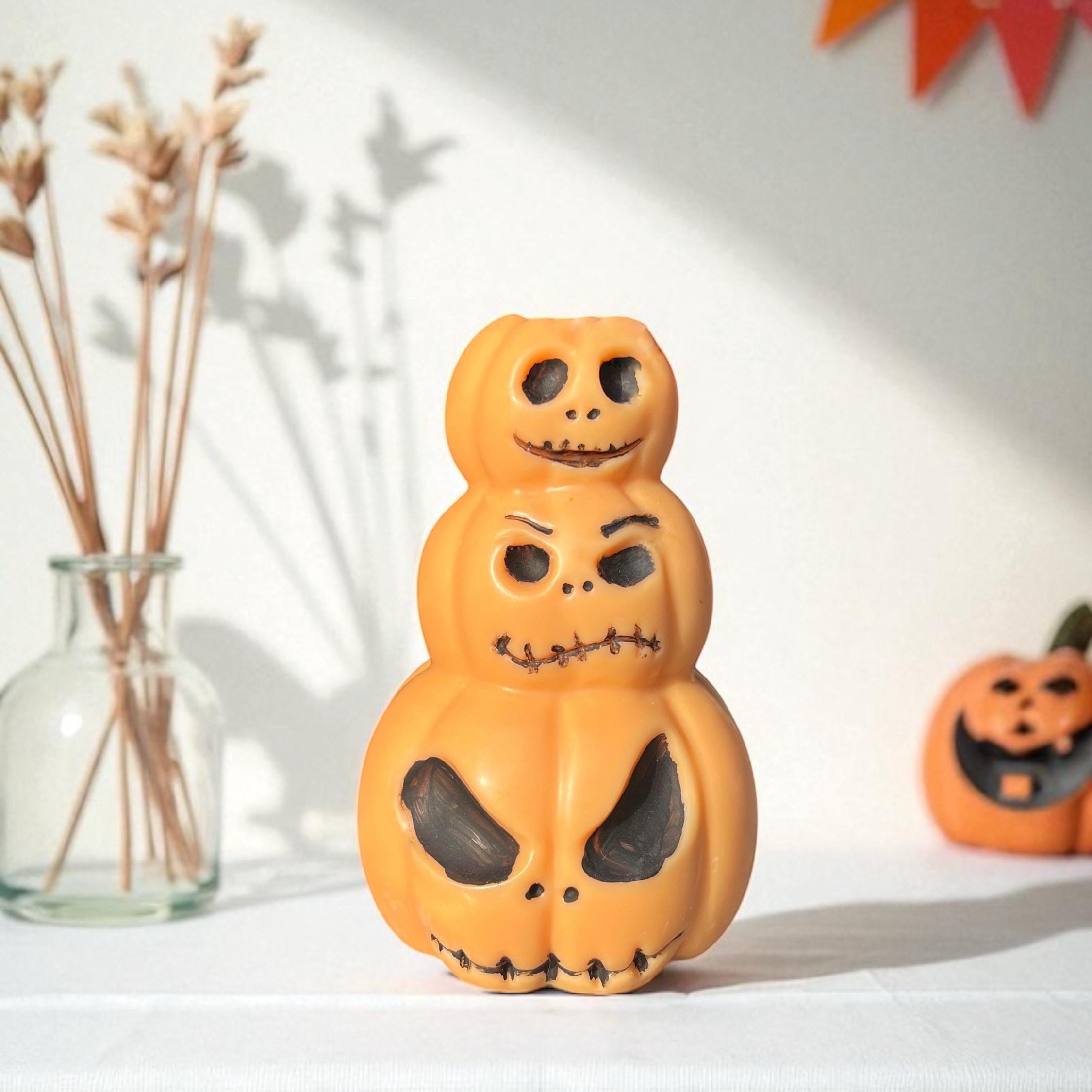 A stacked Halloween decoration featuring three orange jack-o'-lantern shapes with carved black faces, displayed on a white tablecloth with seasonal props in the background.