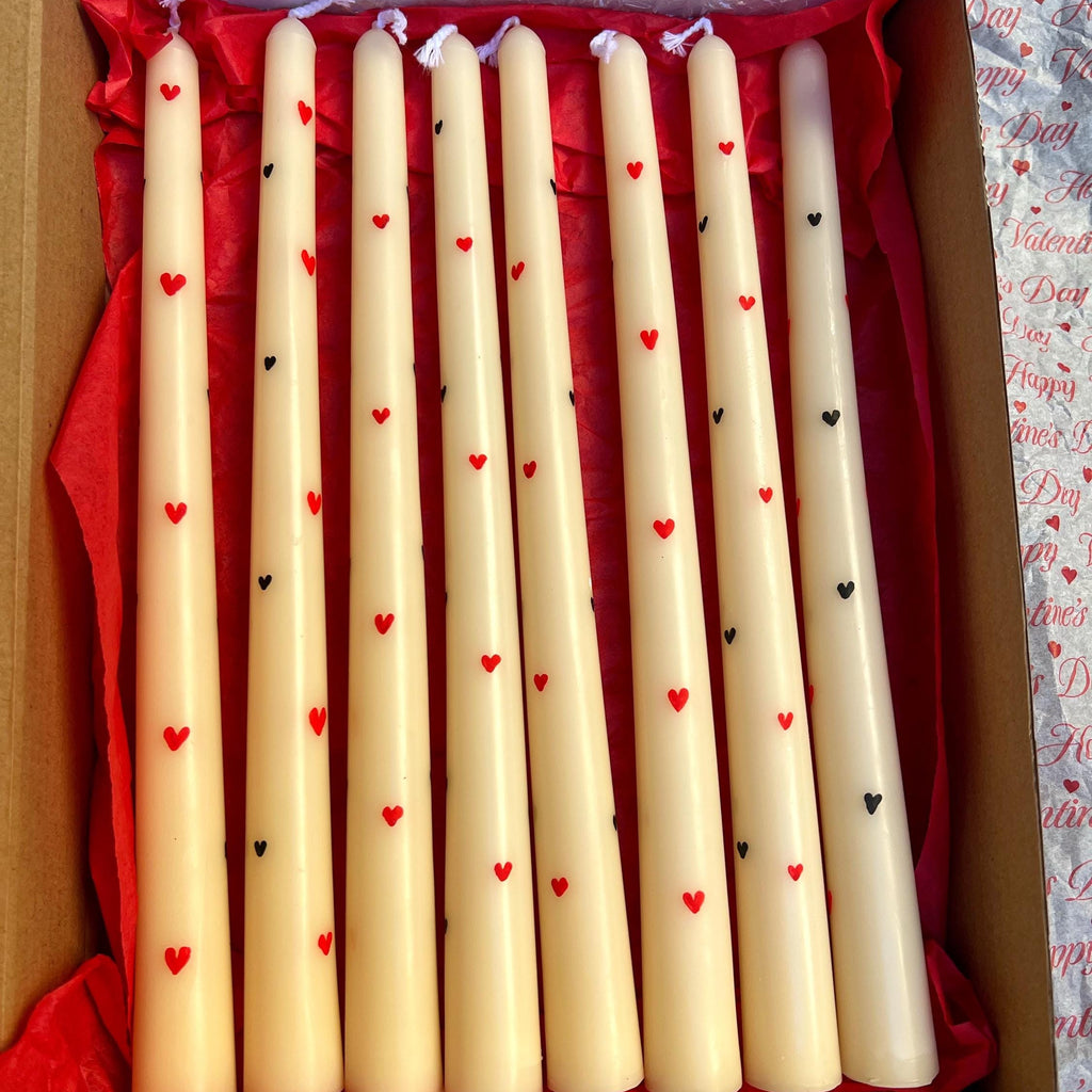 A set of eight cream-colored taper candles with small red and black heart prints, presented in an open gift box lined with red tissue paper.