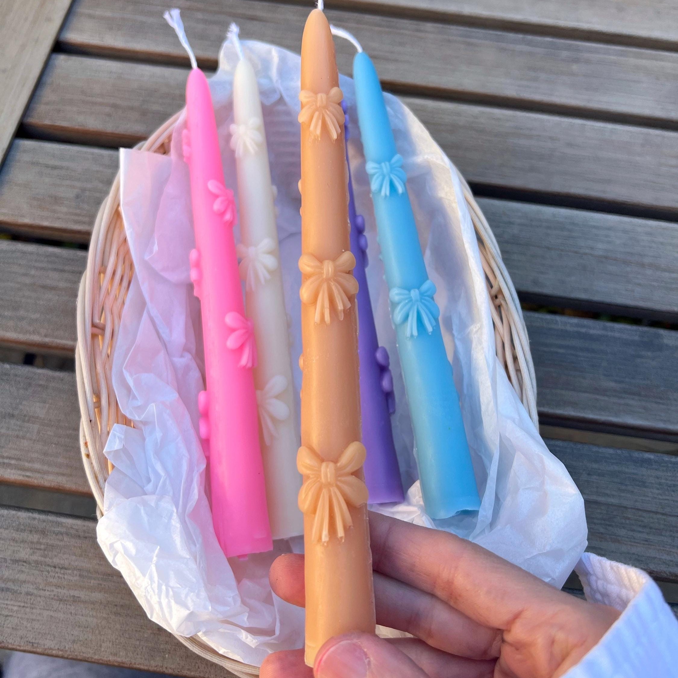 A hand holds a tan taper candle in front of a wicker basket containing four more colored taper candles in pink, white, blue, and purple, set on a wooden table.