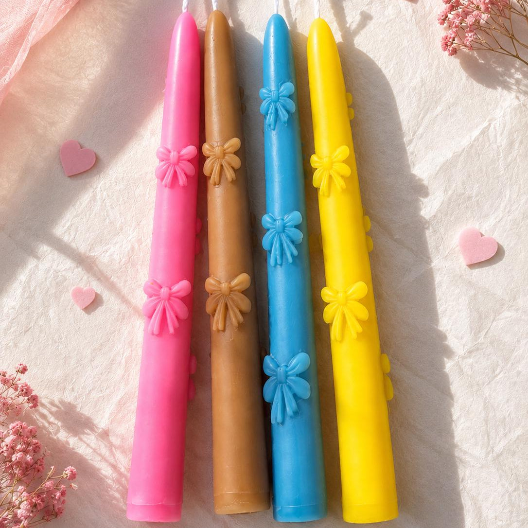Four decorative taper candles in pink, tan, blue, and yellow, each featuring a small bow detail, arranged on a textured white fabric with pink heart confetti and dried flowers.