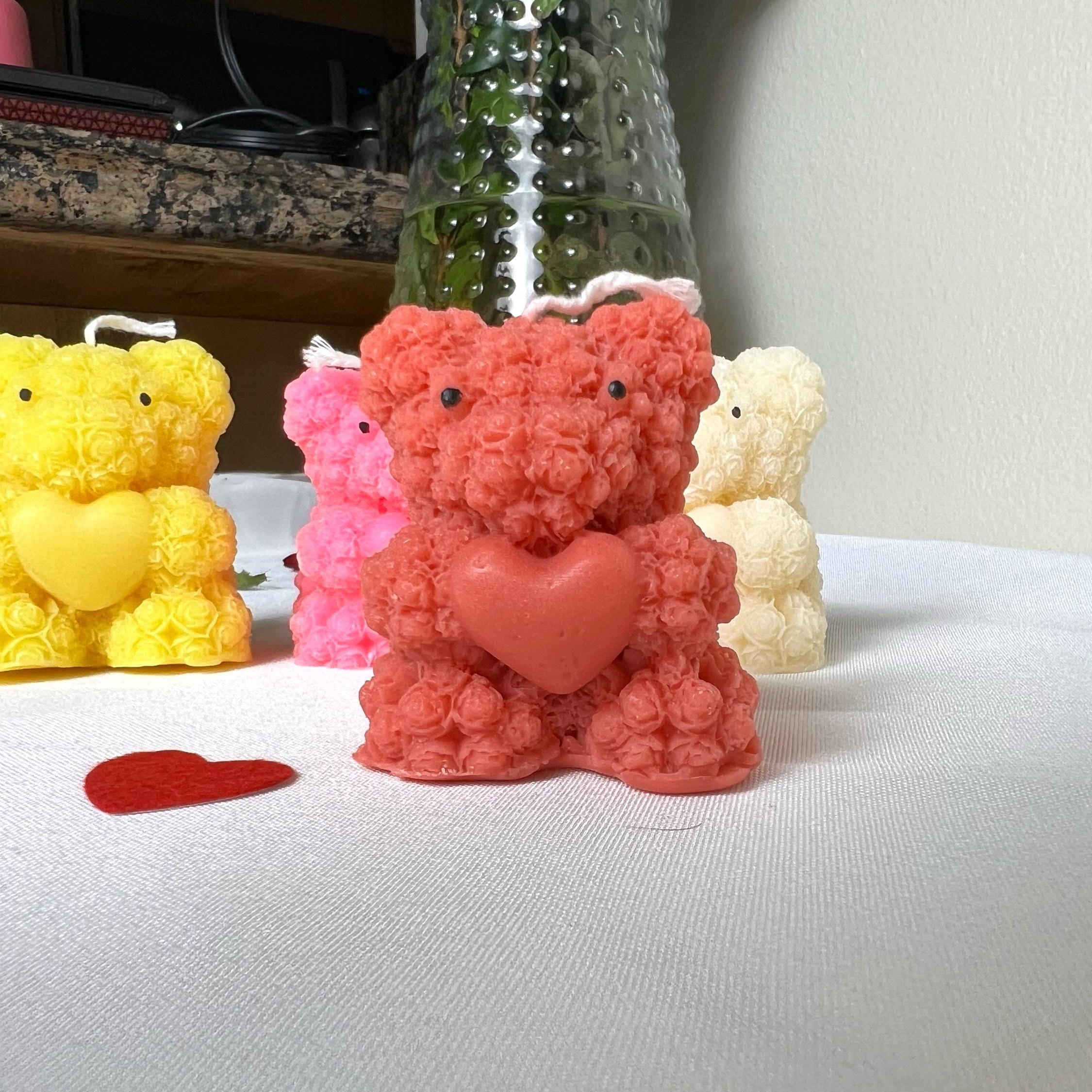 A textured, rosette-style teddy bear candle in coral pink with a matching heart-shaped chest, displayed on a white tablecloth alongside other colored bear candles and a small red heart.