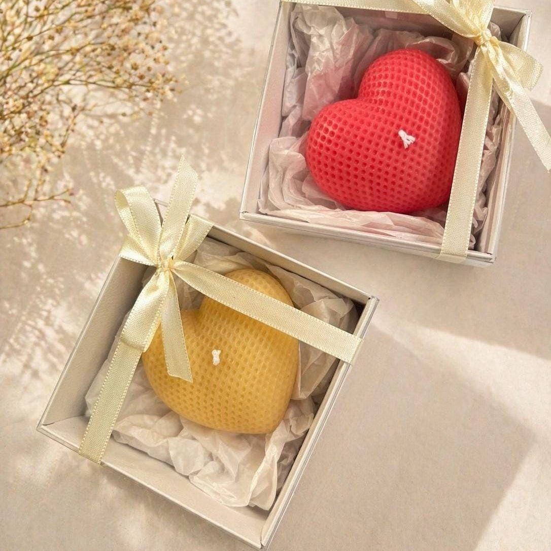 Two heart-shaped textured candles in open gift boxes with cream ribbons. One candle is pale yellow and the other is coral red, both resting on white tissue paper inside light-colored boxes.