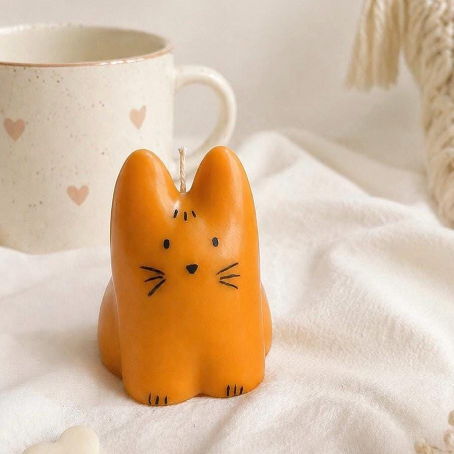 An orange cat-shaped candle with black facial details sits on a white textured fabric surface next to a speckled mug with heart motifs.