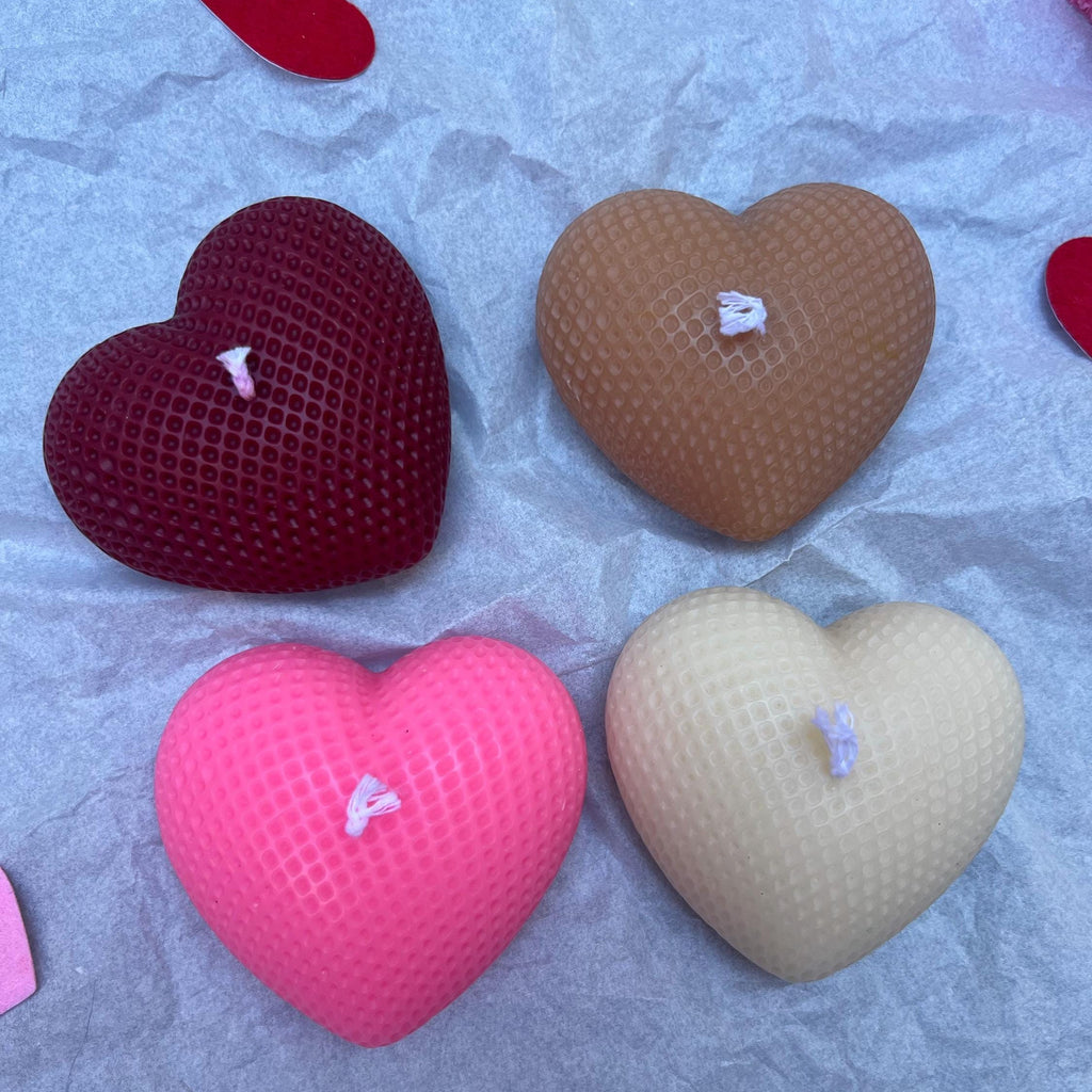 Four heart-shaped textured candles in red, brown, pink, and beige, arranged on a crumpled light blue paper background.