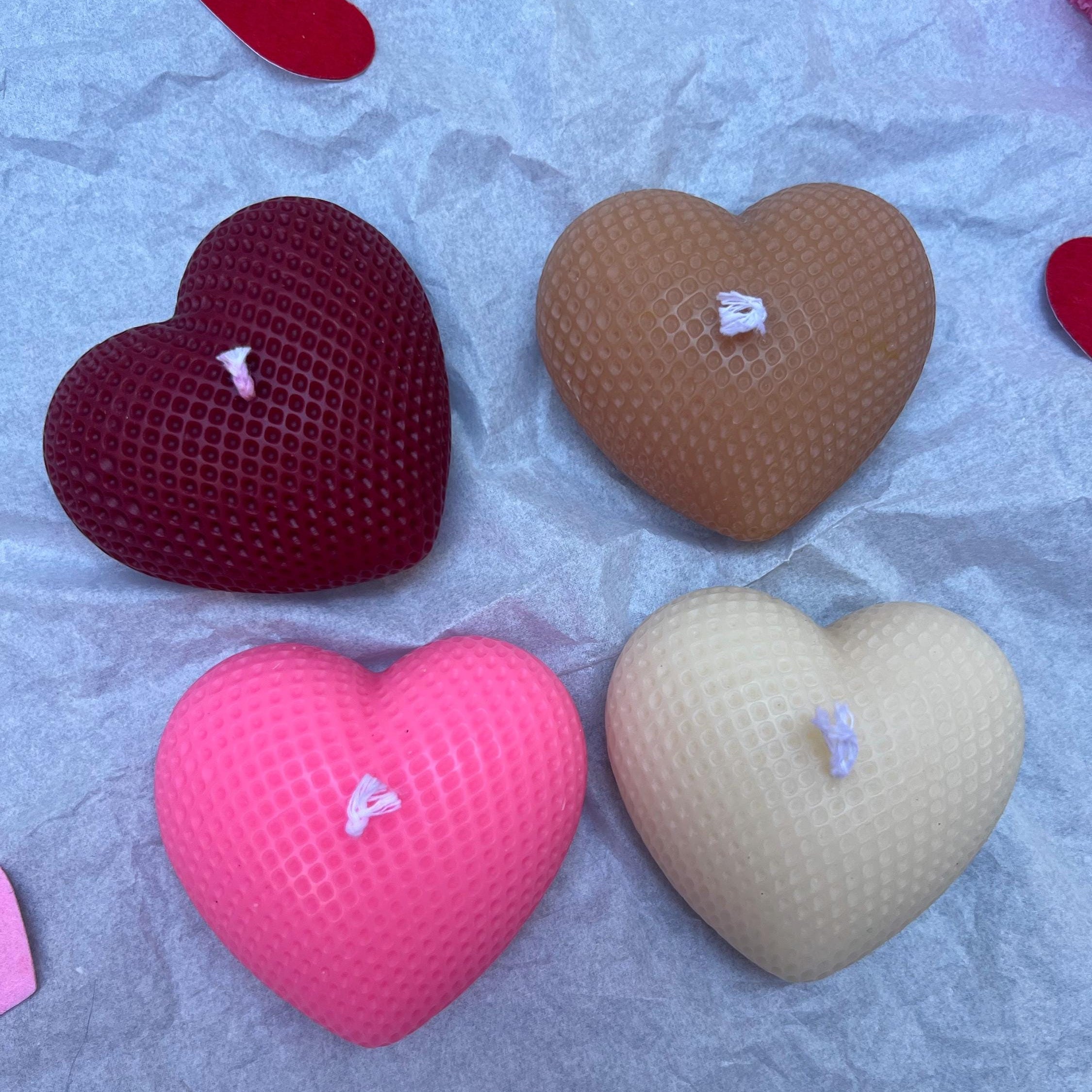 Four heart-shaped textured candles in red, brown, pink, and beige, arranged on a crumpled light blue paper background.