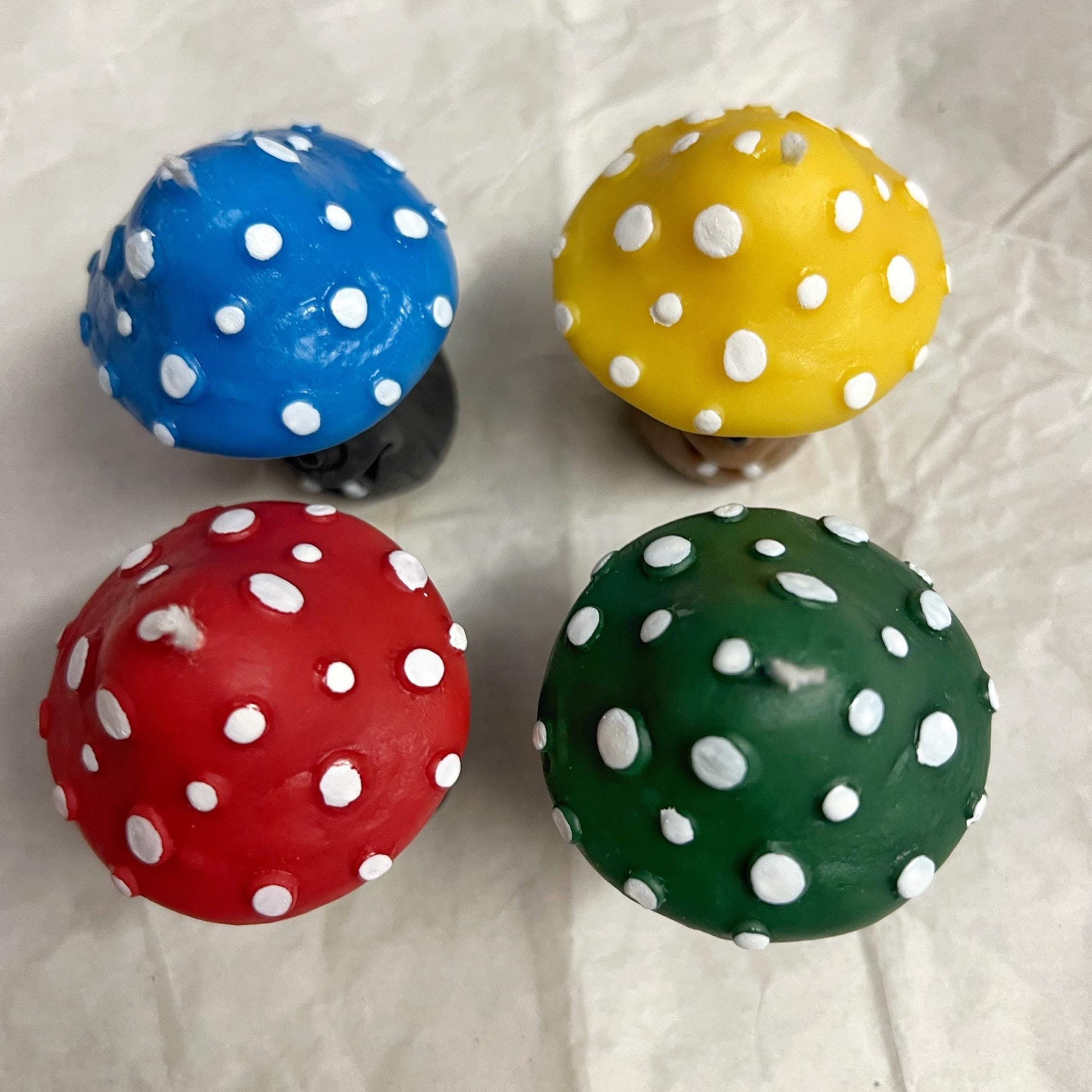 Four mushroom-shaped decorative items arranged in a 2x2 grid on a wrinkled white paper background. The top-left is blue with white spots and a black base, the top-right is yellow with white spots and a brown base, the bottom-left is red with white spots, and the bottom-right is green with white spots.