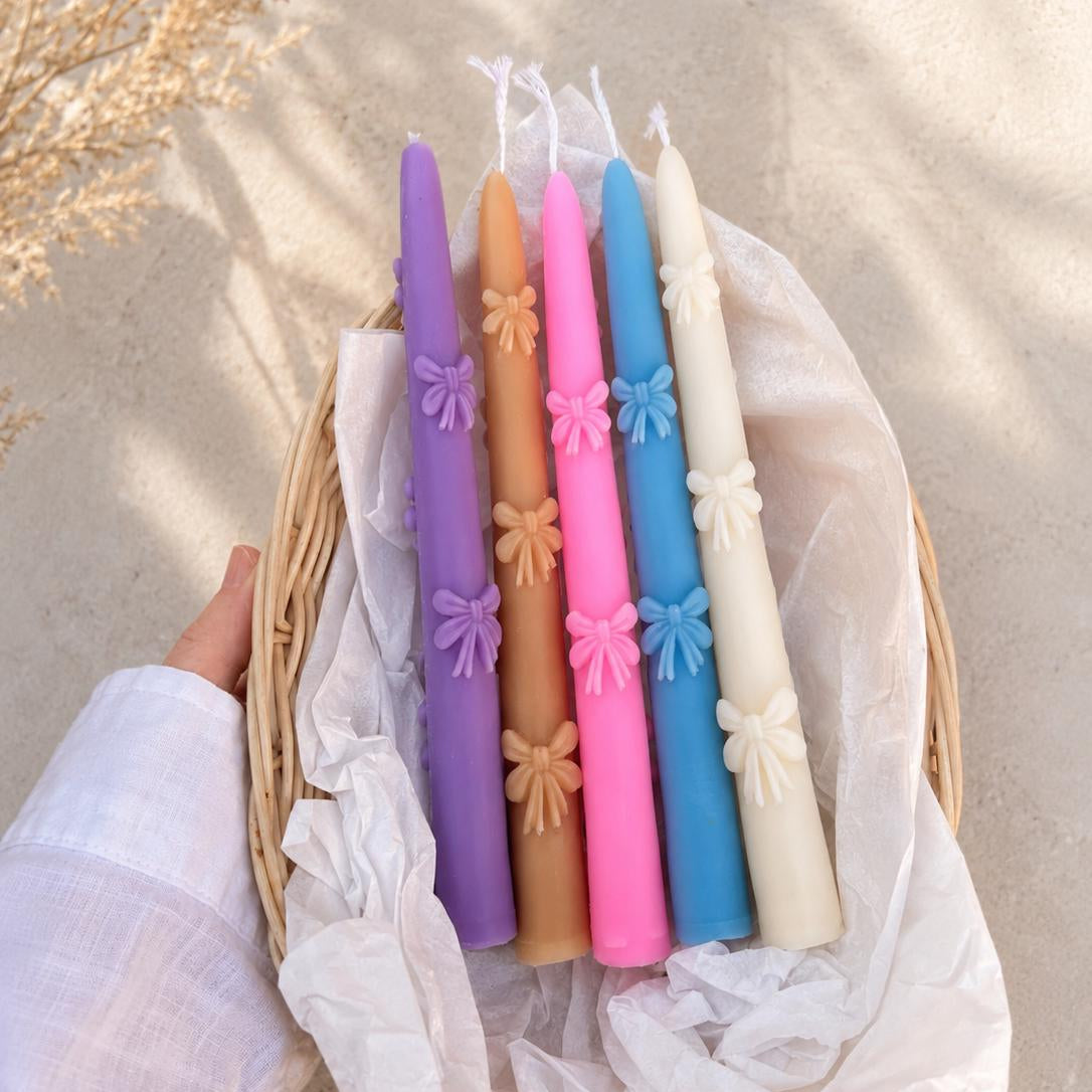 Six colorful taper candles in purple, tan, pink, blue, and cream, decorated with small bow details, arranged in a wicker basket lined with white tissue paper, held by a hand wearing a white sleeve.