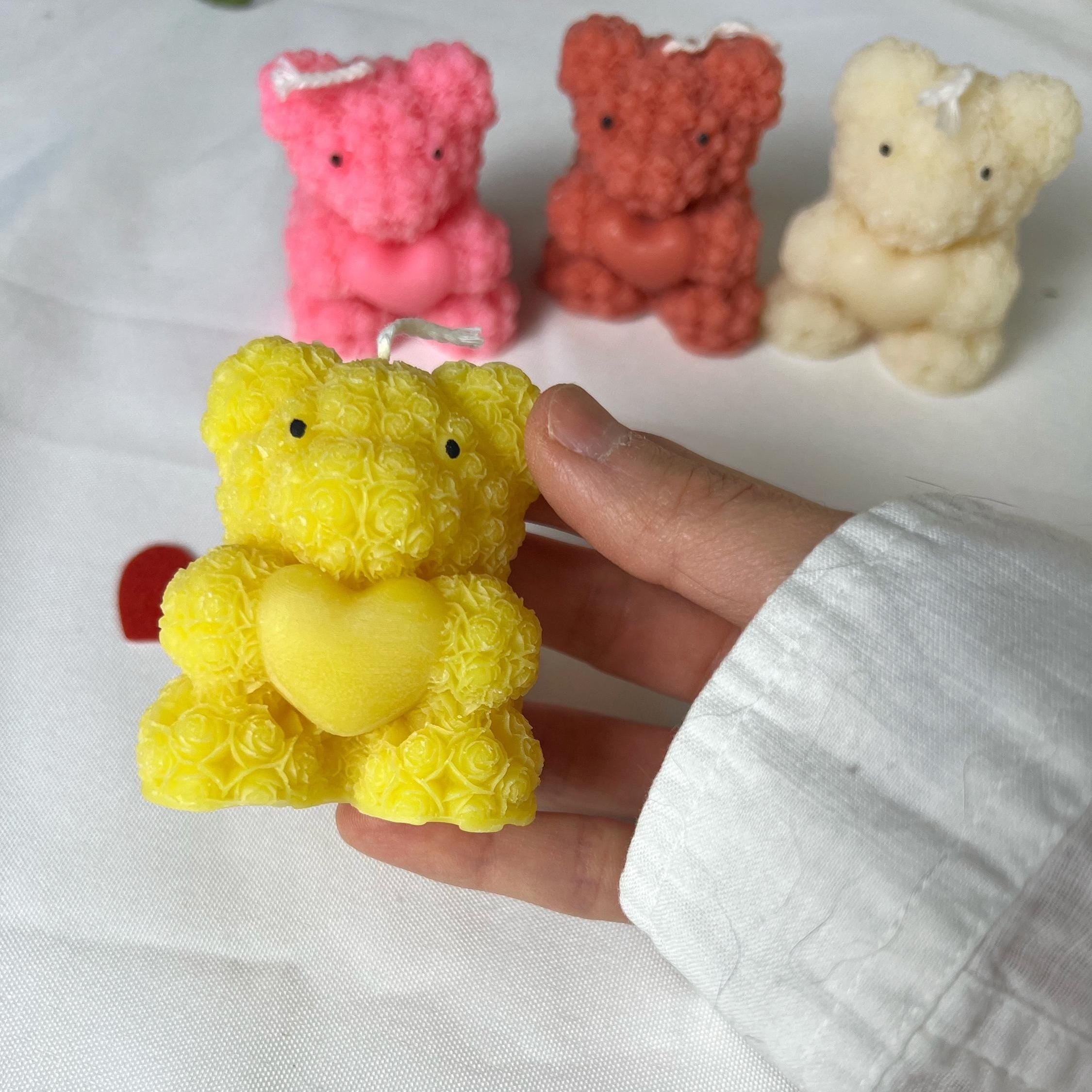 A hand wearing a white long-sleeve shirt holds a yellow textured teddy bear candle with a yellow heart. In the background, three more textured teddy bear candles in pink, orange, and cream are lined up on a white fabric surface.