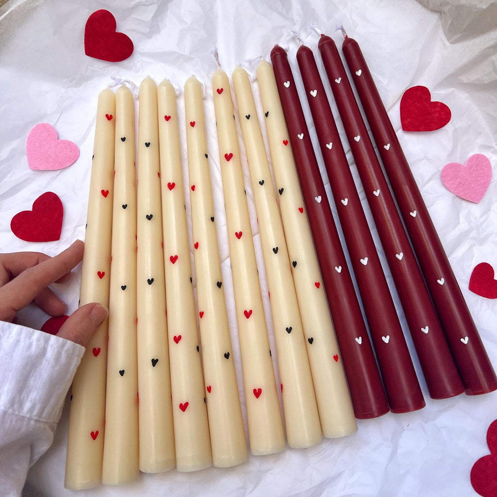 Three sets of long taper candles laid out on white tissue paper: cream candles with small black hearts, dark red candles with small white hearts, and dark red candles with small red hearts. Pink and red felt hearts are scattered around, and a hand in a white sleeve holds one cream candle.