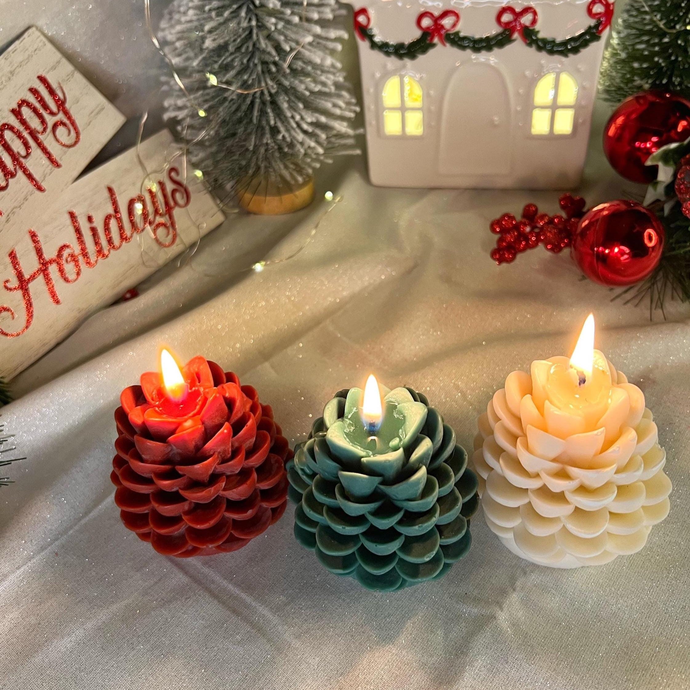 Three pinecone-shaped flameless LED candles in red, teal, and cream are lit on a draped white fabric surface, surrounded by Christmas decorations including a small house, miniature trees, ornaments, and a holiday sign.