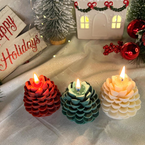 Three pinecone-shaped flameless LED candles in red, teal, and cream are lit on a draped white fabric surface, surrounded by Christmas decorations including a small house, miniature trees, ornaments, and a holiday sign.